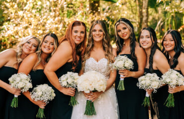 Bride with bridesmaids in black dresses holding bouquets outdoors.