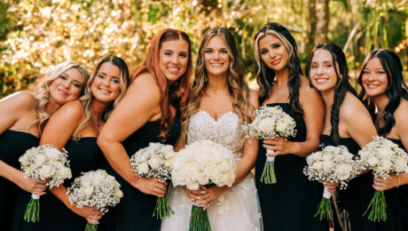 Bride with bridesmaids in black dresses holding bouquets outdoors.